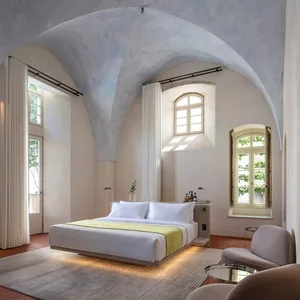 A minimalist bedroom with concrete walls and floor, featuring a large bed, a small armchair, and natural light from multiple 