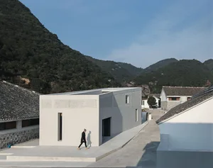 A modern building made of concrete with a person walking on a walkway in front of it, set against a backdrop of green hills a