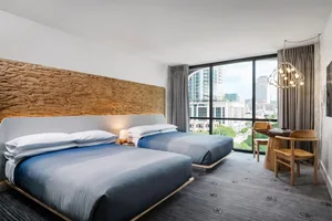 A modern hotel room with two beds, a desk, and a large window providing a view of the city.
