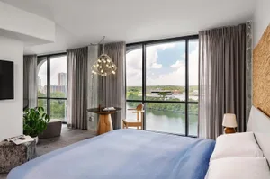 A modern minimalist bedroom with a view of a cityscape and a body of water, featuring a bed with blue bedding, a hanging chan