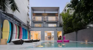 Modern minimalist house with concrete exterior, glass windows, and a pool surrounded by woven lounge chairs and colorful surf