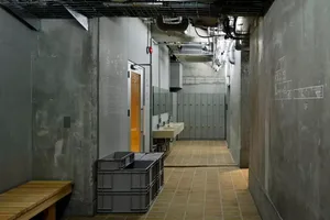A modern public bathroom with concrete walls, a steel sink, and a wooden bench.