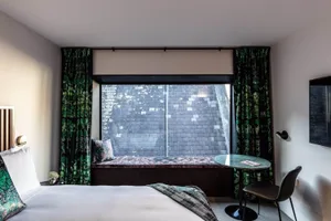 A modern minimalist bedroom featuring a bed with a wooden headboard and white linens, a small table with a black top and chai