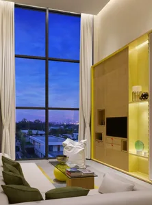 A modern minimalist living room with large windows, white furniture, green accents, and a yellow storage unit with built-in l