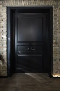 A modern, black wooden door with a panel design, set against a textured brick wall.