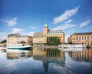 A cityscape featuring stone and glass buildings reflected in calm water.