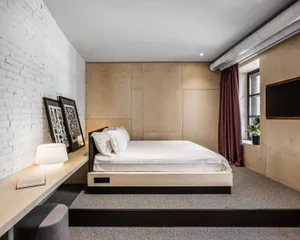 A minimalist bedroom featuring a wooden bed platform with white bedding, a wooden desk with a lamp and framed art, and a wind
