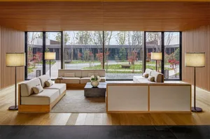 A modern living room with wood paneling, including beige sofas, a glass coffee table, and floor-to-ceiling windows overlookin