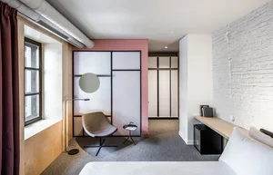 A modern minimalist bedroom with white walls, a wood floor, and a large window with red curtains, featuring a white bed, a wo