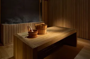 A spacious, minimalist wooden sauna with steam, featuring a wooden table with two wooden buckets and ladles for sauna essenti