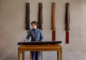A person playing a classical Chinese musical instrument in a room decorated with wooden instruments hanging on the wall.