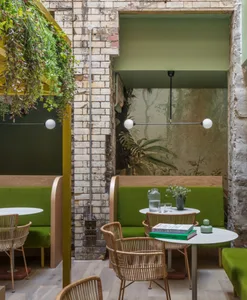 A cozy cafe interior with modern green leather seating, rattan chairs, brick walls, and hanging plants.