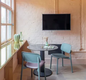A modern industrial cafe setting with a metallic round table and minimalist chairs, featuring a window and a mounted flat-scr