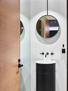 Modern bathroom interior featuring a wooden door, a black pedestal sink with a round mirror above, and a black faucet.