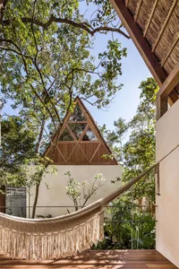A hammock with tassels hanging from a wooden structure with a triangular roof, surrounded by lush greenery.