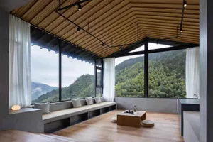 A modern living room with a wooden floor, glass windows overlooking green mountains, and concrete seating areas.