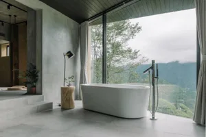 A modern minimalist bathroom with a white freestanding bathtub, grey concrete walls, and a large window overlooking mountains