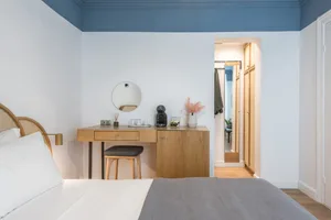 A modern minimalist bedroom featuring a wooden vanity, rattan headboard, and a large bed with white bedding and a grey blanke