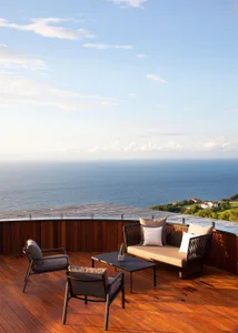 A modern outdoor lounge with a wooden platform, featuring metal chairs and a sofa, overlooking the sea.
