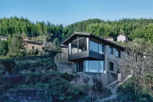 A modern house on a hillside with concrete walls and a glass balcony overlooking the surrounding trees.