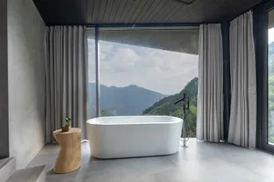 A minimalist bathroom featuring a white bathtub with concrete walls and glass curtains, overlooking mountains and hills.
