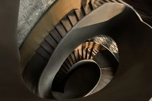 View of a spiral staircase with wooden steps illuminated by natural light, featuring a concrete wall and a rendering backgrou