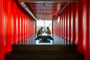 Two individuals seated at a table in a shipping container setting, surrounded by red metal walls.