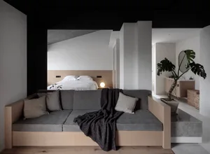 A minimalist living room featuring a floating sofa with gray cushions and a black throw blanket, a concrete headboard bed wit