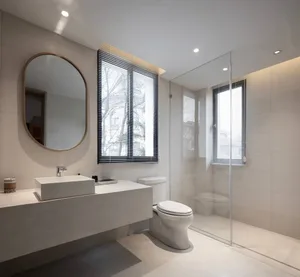 A modern bathroom with white concrete walls, a floating vanity, a large round mirror, and a clear glass shower enclosure.