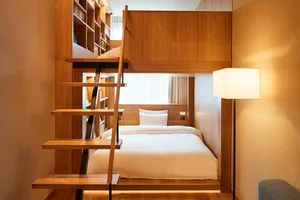 A minimalist bedroom with a wooden bunk bed and stairs, featuring a white pillow and a floor lamp.