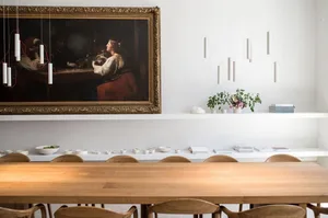 A minimalist dining room with a wooden table and chairs, featuring a hanging light fixture and a framed painting on the wall.