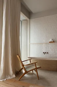 A modern bathroom featuring a tile wall and concrete floor, with a wooden armchair in the foreground and a large shower curta