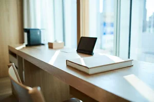 A modern work space featuring a wooden desk with a notebook and tablet, set against a large window.