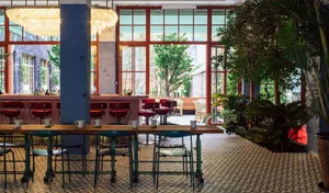 Modern industrial restaurant interior with terrazzo flooring, large windows, green plants, and red chairs.