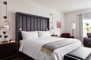 A modern residential bedroom with a gray upholstered headboard, white bedding, and a black leather bench at the foot of the b