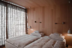 Minimalist bedroom with white bedding, wooden walls, and large window with light-colored curtains.