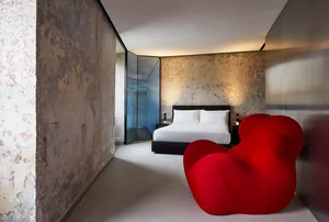Modern bedroom featuring a concrete wall, a red chair, and a neatly made bed with white sheets and pillows.