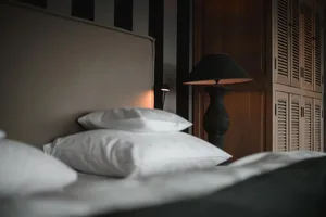 A modern bedroom with a dark headboard and white bedding. The room features a black table lamp, a wooden dresser, and a bed w