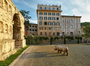 A rhinoceros statue stands on a stone pavement amidst ancient ruins and old buildings.