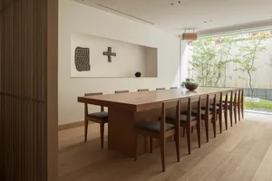 A minimalist dining room featuring a long wooden table surrounded by chairs, with a minimalist wall art piece and a small gar