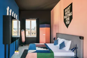 A modern bedroom with pink and blue walls, a patterned blanket, and a poster reading 'Right Here Waiting for You'.