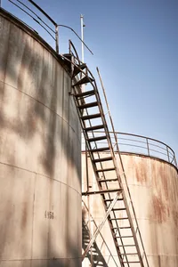 A weathered metal ladder ascending a large cylindrical tank against a clear blue sky.