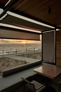 A cozy indoor seating area overlooking the ocean during sunset, featuring wooden furniture and a window view of the beach.