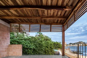 A terrace with a wooden roof overlooking greenery and the sea. The structure features a brick wall and a black metal railing.