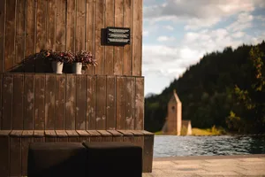 A serene outdoor space with wooden panels, a wooden bench, potted plants, and a view of a church and forested hillside.