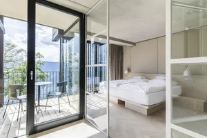 A modern, minimalist bedroom with concrete walls and a glass door leading to a balcony overlooking a scenic view.