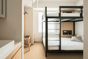 A minimalist hotel bedroom with a black steel bunk bed frame and white bedding, featuring a light wooden stool and a window w