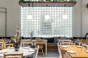 A modern cafe interior with white walls, glass block wall, wood and white tables, and bench seating.