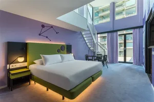 A modern hotel room featuring a green and purple color scheme with a king-sized bed, green bedding, and a purple headboard.