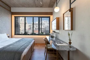 A minimalist bedroom with a concrete wall, wooden floor, and large window overlooking a cityscape.